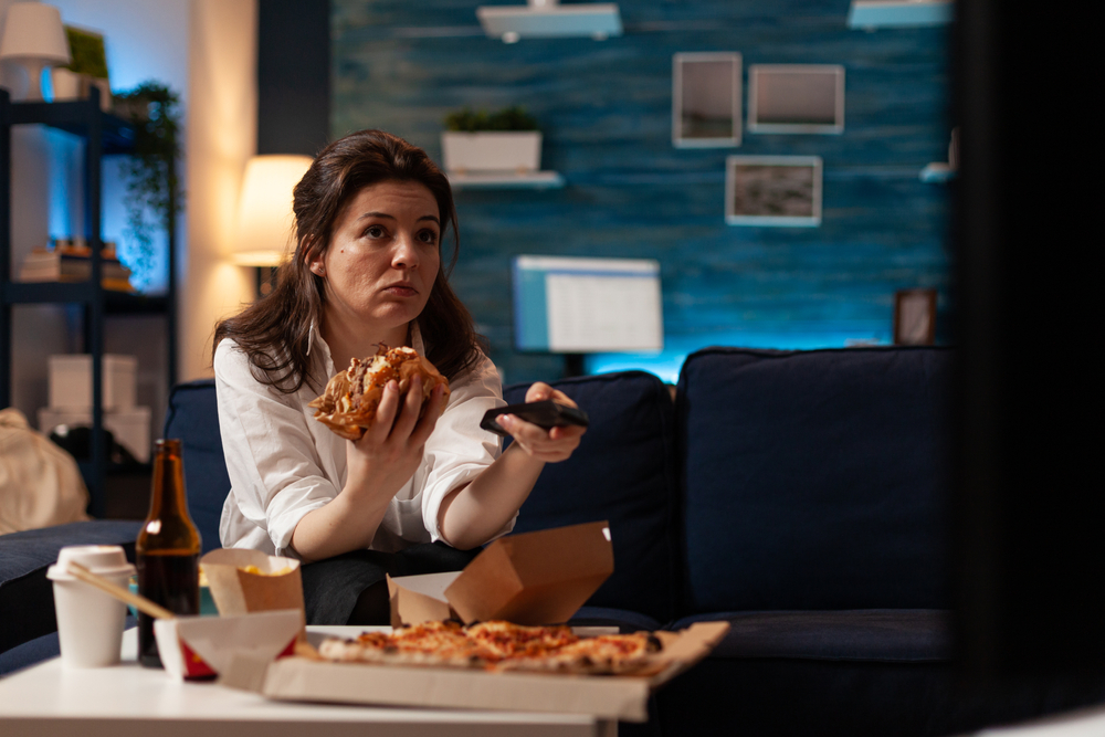 Woman sitting on couch watching TV emotionally eating unhealthy foods consisting of burger , pizza, and french fries accompanied by unhealthy drinks as well.