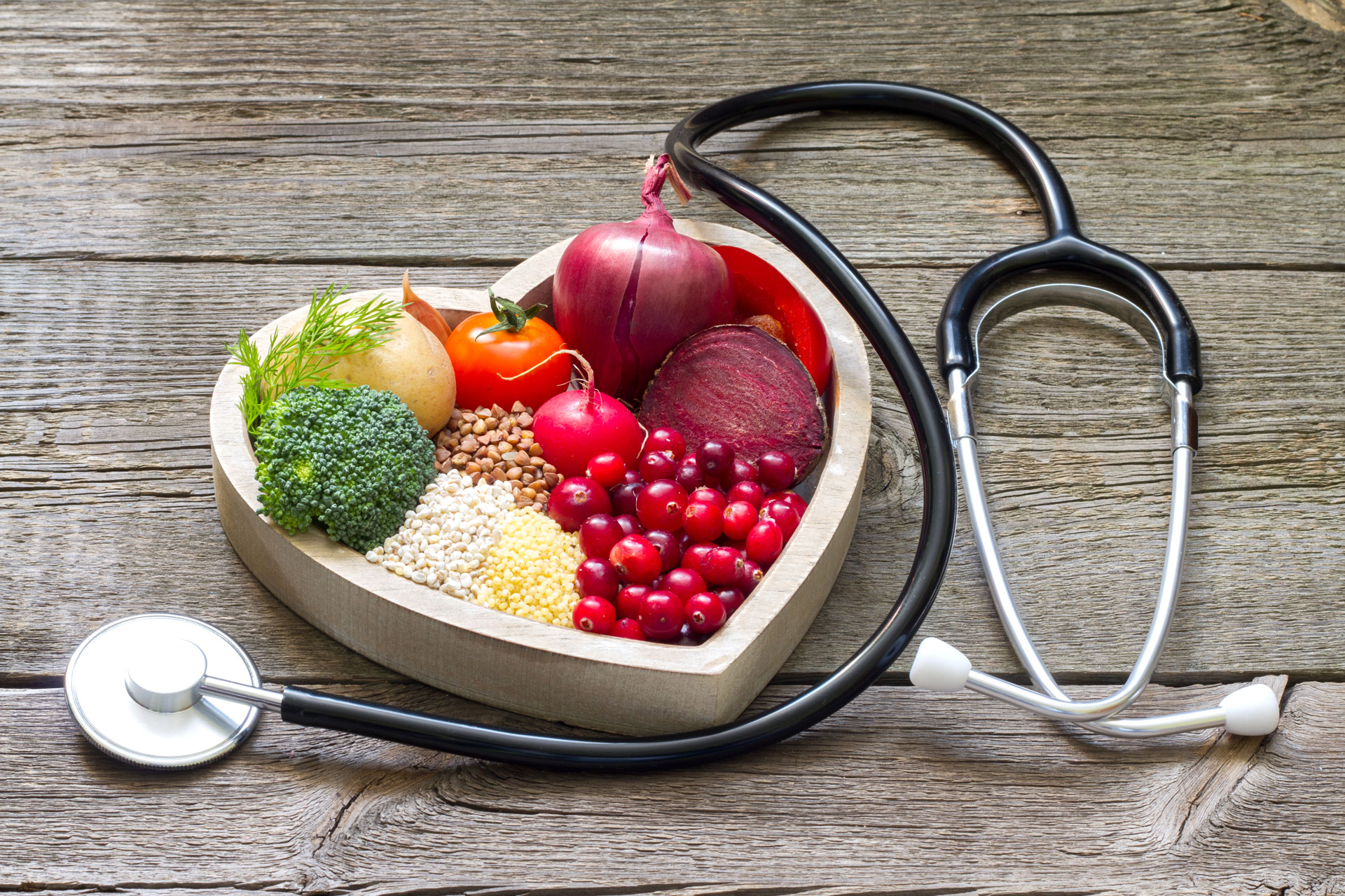 A heart shaped bowl full of heart healthy vegetables on a table that lies next to a stethoscope.