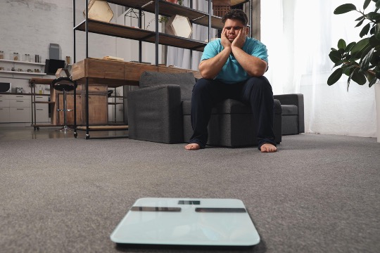 An man sits on the couch and looks at the scale on the floor with a dejected look on his face