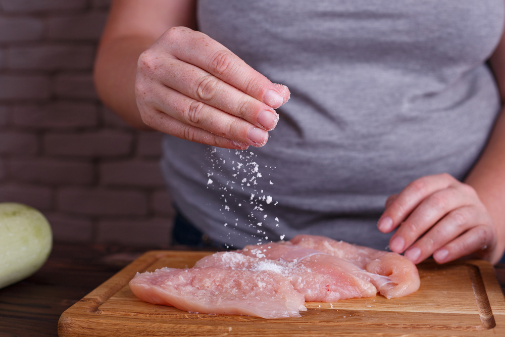 An overweight person sprinkles salt on a raw chicken breast.