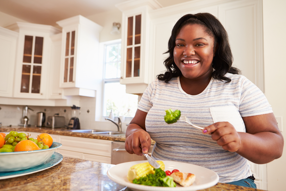 A woman sits at her kitchen counter and eats broccoli and other vegetables high in fiber off a plate as a part of her weight loss journey