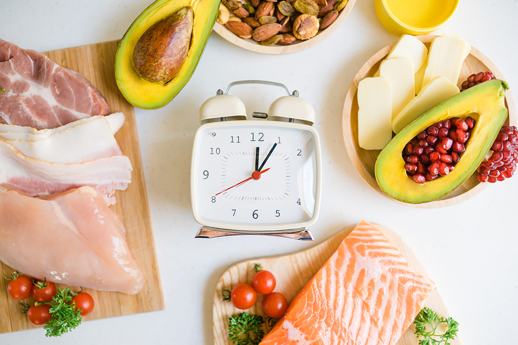 An alarm clock on a counter is surrounded by salmon, avocados, nuts, and other foods.