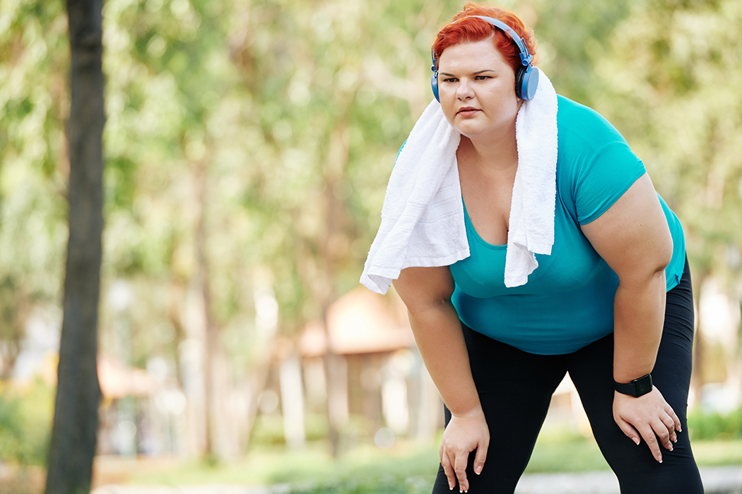 A woman with red hair is bent over with her hands on her knees and is wearing a blue shirt, blue headphones, and a white towel wrapped around her neck. 