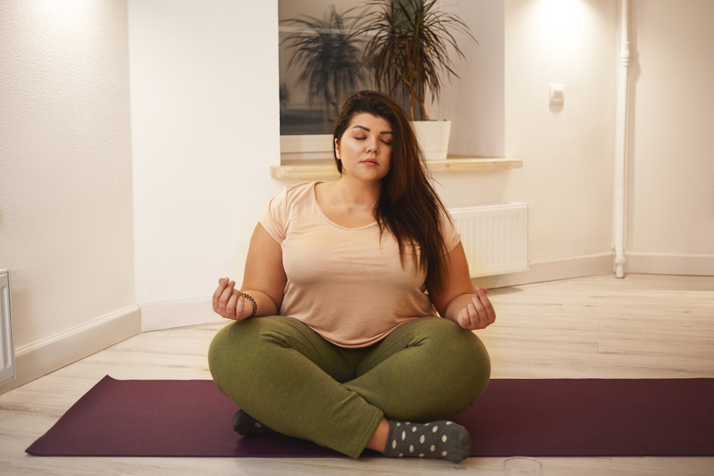A woman wearing a beige shirt and green sweats sits on a yoga mat with her legs crossed and meditates. 