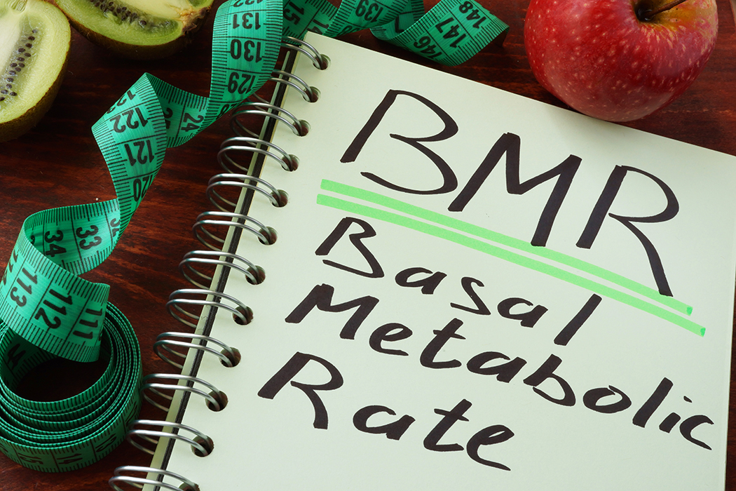 A green notepad with “BMR Basal Metabolic Rate” written in black marker sits on a table beside a green measuring tape, an apple, and a sliced kiwi. 