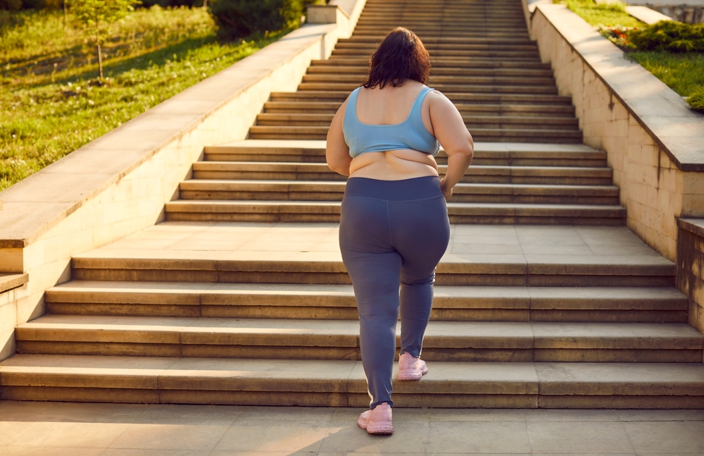 An overweight woman in purple yoga pants, pink running shoes, and a blue tank top begins running up a set of stairs on a sunny day. 