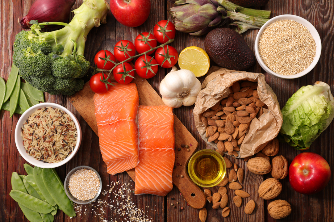 Brown cutting board consisting of healthy foods including salmon, broccoli, tomatoes, lettuce, garlic, lemon, avocado, and asparagus