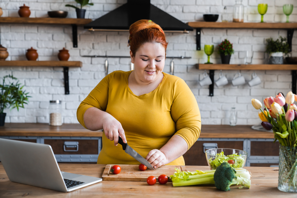 A woman with red hair and a yellow shirt chops vegetables and has her laptop open on the countertop.